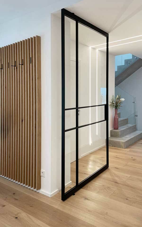 Eine Glastür mit schwarzen Rahmen in einem modernen Raum. Der Fußboden besteht aus hellen Holzbohlen.