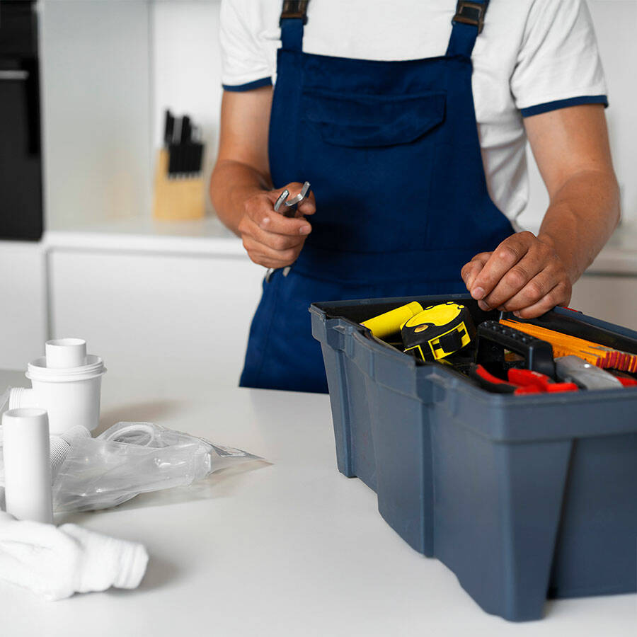 Ein Mann in einem blauen Overall steht vor einem Werkzeugkasten und hält ein Werkzeug in der Hand. Der Kasten enthält verschiedene Werkzeuge.